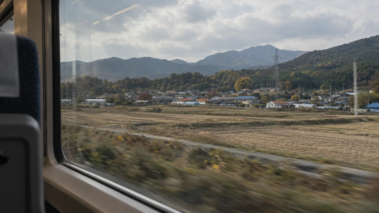 Harvested rice fields, a village and wooded hills seen from a moving train window in South Korea