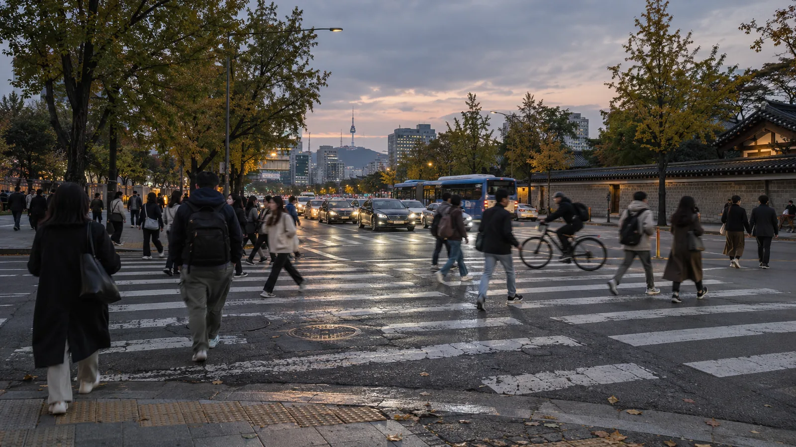 Pedestrians and a cyclist crossing a wide street in central Seoul at dusk, N Seoul Tower in the distance